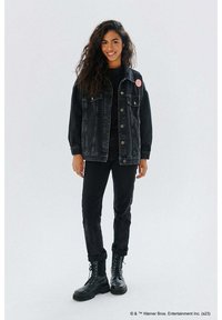 Czarna kurtka dżinsowa z zapięciem na guziki i naszywką, zestawiona z czarnym topem i spodniami. Model nosi czarne sznurowane botki. Gładka faktura przez całość.