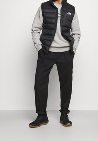 Gilet di piuma nera sopra un felpa grigia, abbinato a pantaloni neri e scarpe scure. Il gilet presenta cuciture orizzontali e un accento con logo.