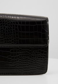 Borsa a mano in pelle nera con finitura lucida, caratterizzata da una texture in coccodrillo e una chiusura a scatto. I bordi sono cuciti con precisione.