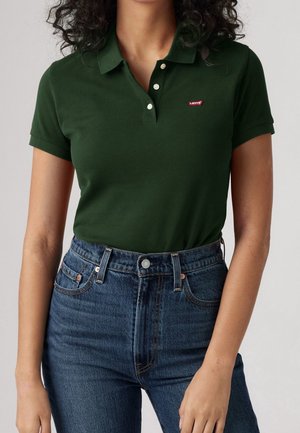Kvinde iført en mørkegrøn polo med korte ærmer med et lille rødt Levi's logo og højttaljede blå denimjeans.