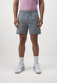 Pantalones cortos deportivos grises con cintura elástica, bolsillos laterales y ribetes de acento negros, combinados con zapatillas blancas y calcetines hasta el tobillo.