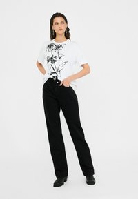 Tricou alb cu imprimeu floral negru, asociat cu pantaloni din denim negri cu talie înaltă. Modelul poartă cizme negre până la gleznă. Design minimalist.