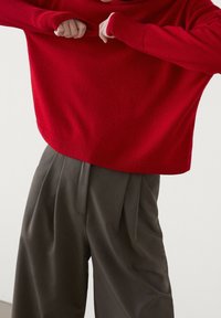 Punainen neulepaita, jossa on rento istuvuus ja ribbireuna, yhdistettynä korkeavyötäröisiin, poimutettuihin tumman vihreisiin housuihin, jotka ovat sileäpintaiset.