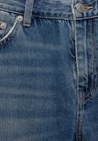 Jean en denim bleu avec un effet délavé, équipé de garnitures en cuivre, un design à cinq poches et des coutures orange le long des coutures.