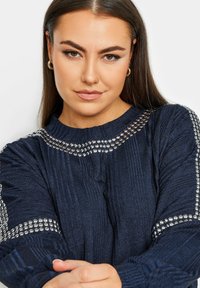 Navyblå tröja med ribbat mönster, prydd med metalliska nitar runt halsringningen och ärmarna. Långa ärmar och avslappnad passform.