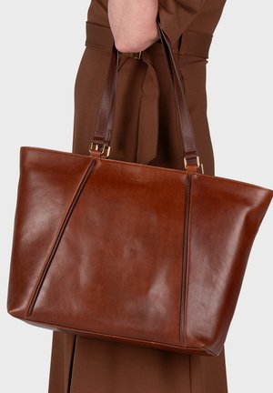 Bolso tote de cuero marrón con asas marrón oscuro, llevado por una persona con un conjunto marrón sobre un fondo claro y liso.