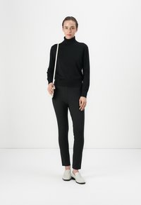 Black turtleneck sweater with ribbed hem, paired with slim-fit black pants. Model holds a white crossbody bag and wears gray slip-on shoes.