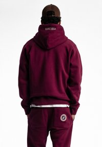 Sweatshirt à capuche bordeaux en tissu doux, présentant un logo à l'arrière, associé à un pantalon assorti avec un détail de poche à patch.