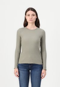 Hosszú ujjú bordázott felső olajzöld színben, kerek nyakkivágással. Az anyag texturált felülettel rendelkezik, és fitted farmernadrággal van párosítva.