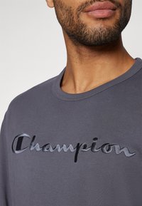 Graues Langarm-T-Shirt mit einem schwarzen gestickten "Champion"-Logo auf der Vorderseite. Weicher Stoff mit einem klassischen Rundhalsausschnitt.