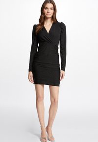 Robe noire à manches longues avec un décolleté en V et des détails froncés. Fabriquée en tissu texturé avec un léger éclat. Silhouette ajustée, longueur au genou. Sandales argentées.
