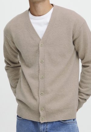 Mann trägt einen beigen, geknöpften Cardigan über einem weißen T-Shirt, die Hände hinter dem Rücken, kombiniert mit hellblauen Jeans.