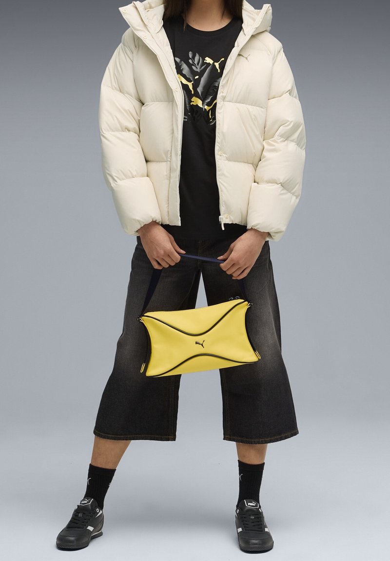 Personne portant une doudoune blanche, un t-shirt noir Puma, un jean large noir, des chaussettes et chaussures noires, tenant un sac à main jaune Puma par sa bandoulière.