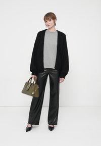 Fekete kötött kardigán egy szürke felsőn, fényes fekete nadrággal párosítva. Olíva zöld kézitáskát tart, csillag akcentussal. Hegyes fekete cipők.