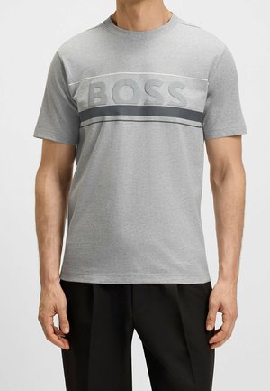 Mężczyzna ubrany w jasnoszary T-shirt z krótkim rękawem z wytłoczonym tekstem "BOSS" i czarne spodnie na jednolitym tle.