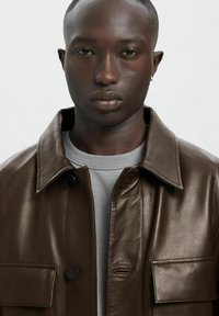 Rjava usnjena jakna z ovratnikom, dvema sprednjima žepoma in zapiranjem na gumbe, nošena preko sive majice z okroglim izrezom. Gladek videz.