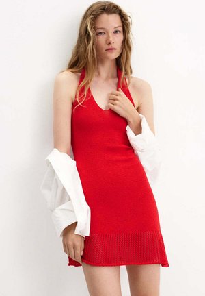 Mujer joven con el pelo largo, vestida con un vestido de punto rojo sin mangas y una camisa blanca sobre los brazos, sobre un fondo liso.