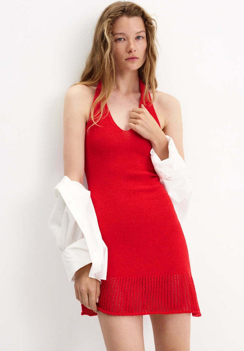 Jeune femme aux longs cheveux portant une robe tricotée rouge sans manches, une chemise blanche drapée sur les bras, devant un fond uni.