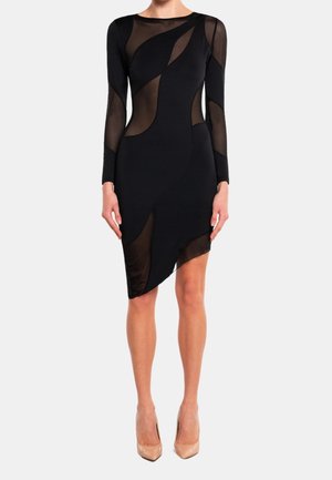 Frau trägt ein eng anliegendes schwarzes Kleid mit transparenten Mesh-Einsätzen und langen Ärmeln, kombiniert mit hautfarbenen spitzen High Heels.