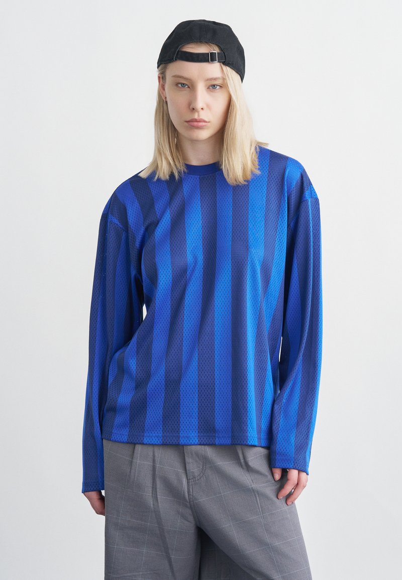 Νεαρός με ξανθά μαλλιά μέχρι τους ώμους, φοράει μαύρο καπέλο προς τα πίσω, μπλε ριγέ πουκάμισο με μακριά μανίκια και γκρι παντελόνι, με ουδέτερη έκφραση.