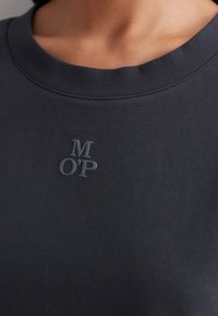 Mörkblå bomullströja med rund hals, med en liten, texturerad grå logotyp "M OP" på övre vänstra sidan.