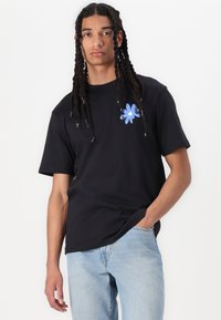 Sort bomulds t-shirt med et blåt blomsterdesign på venstre side, rund hals, korte ærmer og afslappet pasform, parret med lyseblå jeans.
