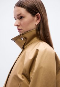 Αδιάβροχο μπουφάν χακί με ψηλό γιακά, κουμπιά με πατητό κούμπωμα και λεία, ματ υφή. Το μοντέλο εμφανίζεται σε προφίλ με ουδέτερο φόντο.