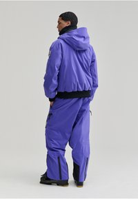 Chaqueta aislante morada con capucha, con dobladillo y puños elásticos negros, acompañada de pantalones a juego. Incluye detalles y cremalleras negras.