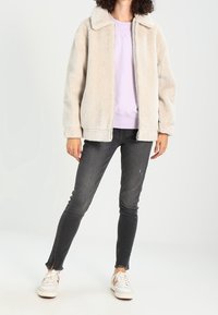 Gräddfärgad fluffig jacka med krage, bärs över en ljus lila sweatshirt, ihop med mörka skinny jeans och vita sneakers.