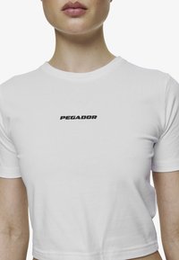 Tricou alb, cu mâneci scurte, scurt, confecționat din bumbac. Prezintă un logo negru "PEGADOR" imprimat pe piept. Textură netedă.