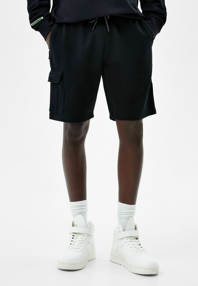 Shorts cargo noirs en coton avec poches latérales et taille à cordon, associés à des baskets montantes blanches et des chaussettes côtelées.