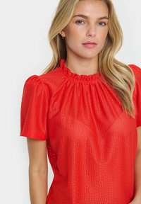 Blusa rossa a maniche corte con colletto arricciato e motivo a ritagli geometrici. Il tessuto è leggero e ha una consistenza morbida.