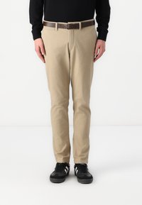 Beige Chinos mit geradem Schnitt, strukturiertem Stoff und Vordertaschen, kombiniert mit einem braunen Gürtel und schwarzen Sneakern mit weißen Streifen.