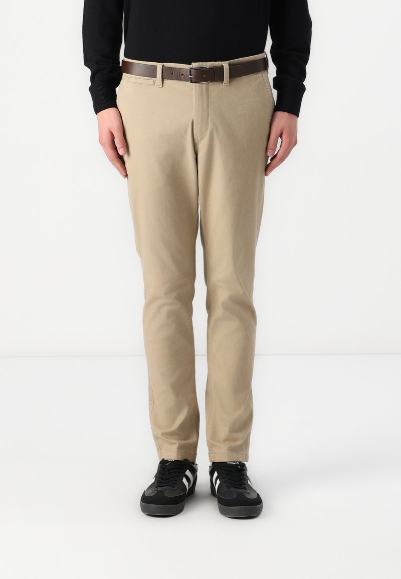 Beige Chinos mit geradem Schnitt, strukturiertem Stoff und Vordertaschen, kombiniert mit einem braunen Gürtel und schwarzen Sneakern mit weißen Streifen.