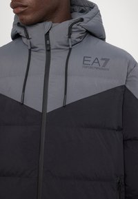 Giacca a vento imbottita con cappuccio, caratterizzata da un design a blocchi di colore in grigio e nero. Il materiale sembra liscio, con cuciture visibili e dettagli del logo.