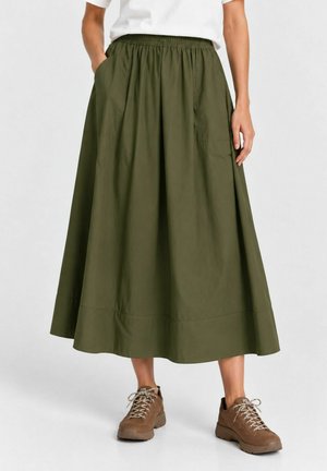 Femme portant une jupe midi vert olive avec des poches et des chaussures de randonnée marron à lacets, debout avec une main dans une poche.