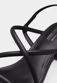 Μαύρο σανδάλι με λεπτά λουράκια από λείο δέρμα που διασταυρώνονται πάνω στο πέλμα, εμφανιζόμενο σε κοντινή λήψη με φόντο λευκό.