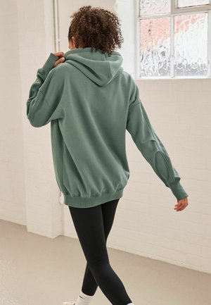 Persona con capelli ricci che indossa un ampio hoodie verde e leggins neri, mentre cammina all'interno vicino a una finestra ghiacciata.