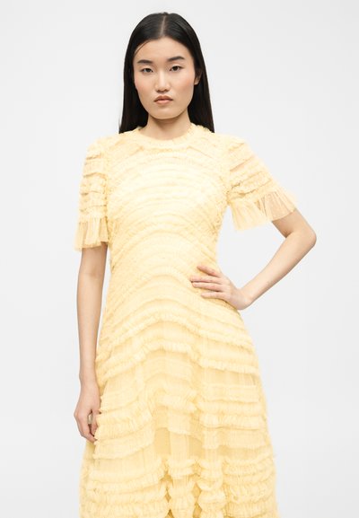 Γυναίκα με μακριά μαύρα μαλλιά που φοράει ανοιχτόκίτρινο φόρεμα με υφή και κοντά μανίκια, στέκεται με το ένα χέρι στη μέση της μπροστά σε απλό φόντο.