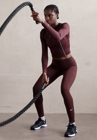 Maroon urheilullinen paita ja legginsit, joissa on valkoinen yksityiskohta, pitää mustasta taistelunarusta kiinni neutraalilla taustalla. Mustat lenkkarit.