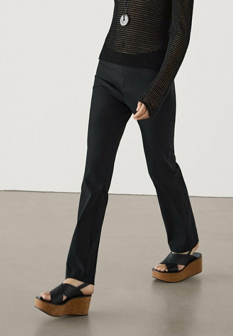 Personne portant un haut en maille noire à manches longues, un pantalon noir ajusté et des sandales compensées noires avec semelles en liège marchant sur un sol uni.