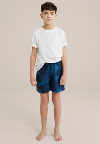 Balta trumpa rankovė t-shirt su kišene, derinama su mėlynomis raštuotomis maudymosi šortais, turinčiais elastingą raištelį ir išlinkusį apatinį kraštą.