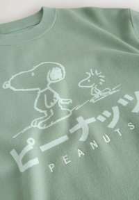 Sweat-shirt vert clair avec des dessins en ligne blanche de Snoopy et Woodstock, accompagné de texte en japonais et de "PEANUTS" en dessous.