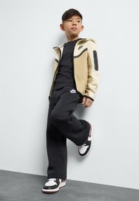 Nike kreminė ir juoda zip-up striukė, juodas marškinėliai, juodos sportinės kelnės su logotipu ir juodai/baltos sportinės avalynės. Lygus audinys, sportiškas telpa.