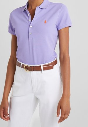 Persona con camisa tipo polo de manga corta color púrpura claro con logo naranja y pantalones blancos sujetos con un cinturón de cuero marrón con hebilla dorada.