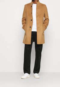 Cappotto in misto lana marrone con colletto alto, chiusura frontale a bottoni e tasche laterali, abbinato a pantaloni in corduroy neri e sneakers bianche.
