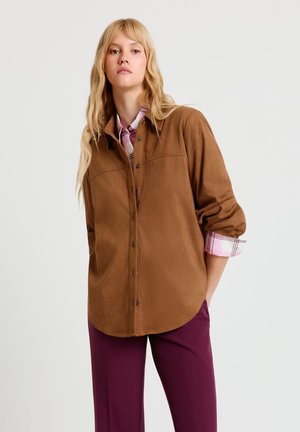 Braunes Wildlederhemd mit Knopfleiste und zwei Brusttaschen, mit hochgekrempelten Ärmeln und einem pink karierten Detail an den Bündchen. Kombiniert mit dunkelvioletten Hosen.