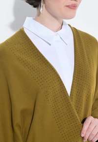 Olivgrüner Pullover mit V-Ausschnitt, verziert mit kleinen metallischen Punktschmuckelementen. Über einem weißen Kragenhemd getragen, was die kontrastierenden Farben betont.
