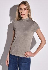 Short-sleeve, high-neck top in metallic beige fabric with a ribbed texture. Worn with dark blue jeans, showcasing a fitted silhouette.