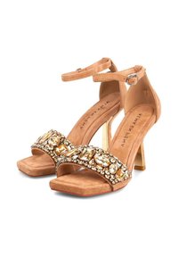 Zapatos de tacón alto en color nude, con la punta abierta y una tira adornada con joyas que presenta acentos dorados y de cristal. Textura de ante suave, tacón delgado y tira para el tobillo.